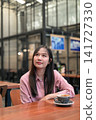 Thoughtful Asian Woman Looking Up while Sitting in a Cafe with Coffee 141727330