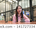 Smiling Asian Woman Sitting with a Cup of Coffee in a Modern Cafe 141727334