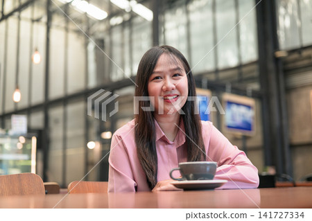 Smiling Asian Woman Sitting with a Cup of Coffee in a Modern Cafe Smiling Asian Woman Sitting with a Cup of Coffee in a Modern Cafe 141727334