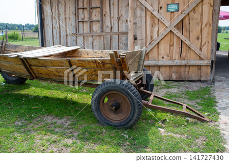 Rustic wooden wheelbarrow and cart beside weathered barn wall, farm equipment resting outdoors on grass near rural fields with wooden planks and large metal wheel Rustic wooden wheelbarrow and cart beside weathered barn wall, farm equipment resting outdoors on grass near rural fields with wooden planks and large metal wheel 141727430