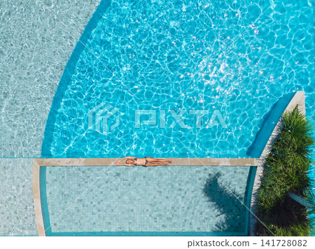 Young woman relax at the edge of clear blue pool during sunny summer vacation. This aerial view captures family,  friends, fun, and recreation at resort, relaxation and joyful outdoor moments. 141728082