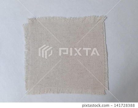 A square swatch of light beige, loose coarse woven fabric with fringed edges, placed on a plain white background. A square swatch of light beige, loose coarse woven fabric with fringed edges, placed on a plain white background. 141728388