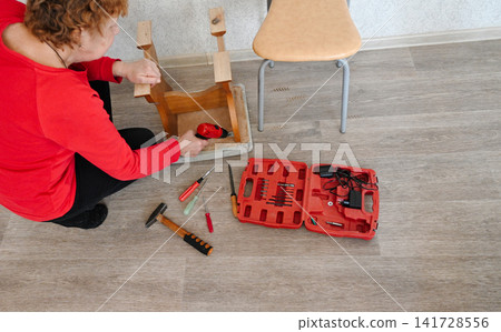 Elderly woman in orange casual clothes assembles wooden chair with her own hands. 141728556