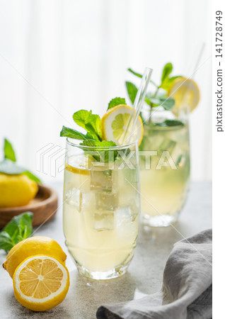 A summer drink with lemon, mint, and ice in a two glasses against a sunny window background. A summer drink with lemon, mint, and ice in a two glasses against a sunny window background. 141728749