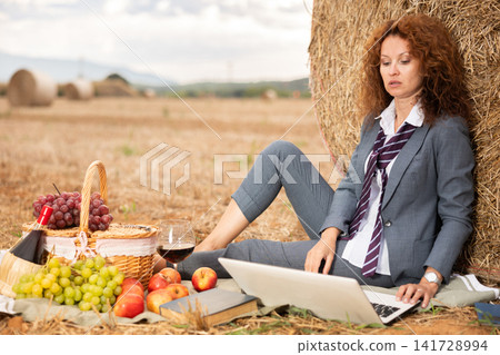 Portrait of woman in office clothes with laptop on picnic at farm field Portrait of woman in office clothes with laptop on picnic at farm field 141728994