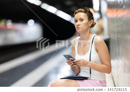 Adult woman sits at subway station with phone Adult woman sits at subway station with phone 141729215