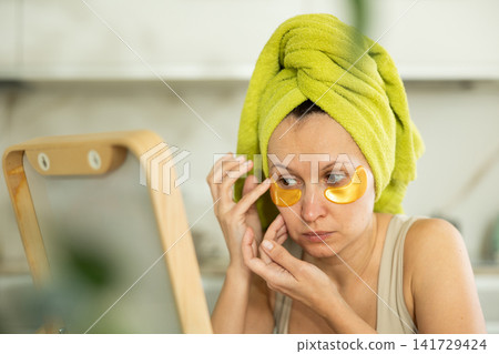 Middle-aged woman applying eye patch mask on her face sitting in front of the mirror Middle-aged woman applying eye patch mask on her face sitting in front of the mirror 141729424