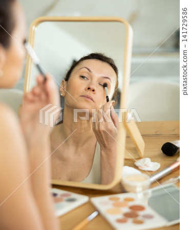 Middle-aged woman using brush during eye makeup sitting in front of the mirror Middle-aged woman using brush during eye makeup sitting in front of the mirror 141729586