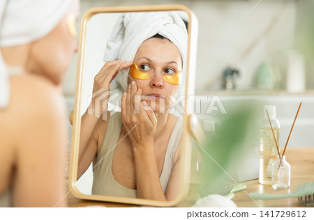 Woman in towel turban applying golden collagen eye patches Woman in towel turban applying golden collagen eye patches 141729612