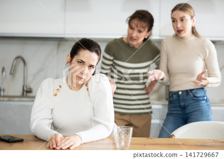 Upset middle-aged woman sitting at the kitchen table with her back to women quarreling to her Upset middle-aged woman sitting at the kitchen table with her back to women quarreling to her 141729667