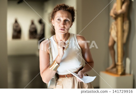Interested young woman looking at collection of medieval sculptures and objects in museum Interested young woman looking at collection of medieval sculptures and objects in museum 141729916