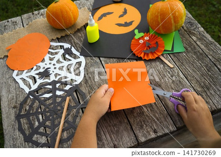 Close up of a child using purple safety scissors to cut out shapes from a bright orange paper sheet Close up of a child using purple safety scissors to cut out shapes from a bright orange paper sheet 141730269