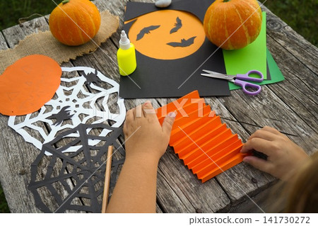 Close up of kids hands folding orange paper like an accordion to create a volumetric Halloween pumpkin craft Close up of kids hands folding orange paper like an accordion to create a volumetric Halloween pumpkin craft 141730272