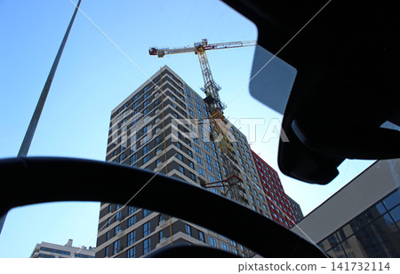 High Rise Construction Tower Crane Near New Building Is Visible Through Car Windshield. Arm Of Construction Crane Over Building At Construction Site High Rise Construction Tower Crane Near New Building Is Visible Through Car Windshield. Arm Of Construction Crane Over Building At Construction Site 141732114