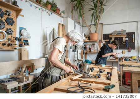 Concentrated master technician setting up handheld wood router instrument for work in workshop. Concentrated master technician setting up handheld wood router instrument for work in workshop. 141732807