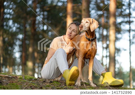 Relaxed woman and Vizsla dog enjoying day out in woodlands together, sit on meadow observing forest. Relaxed woman and Vizsla dog enjoying day out in woodlands together, sit on meadow observing forest. 141732890