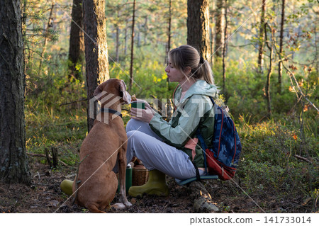 Positive woman backpacker hiking with dog in forest, sitting on log relaxing and eating snacks. 141733014