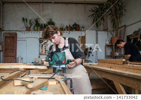 Teamwork and craftsmanship in restoration workshop, woman crafter processing board with wood router. Teamwork and craftsmanship in restoration workshop, woman crafter processing board with wood router. 141733042
