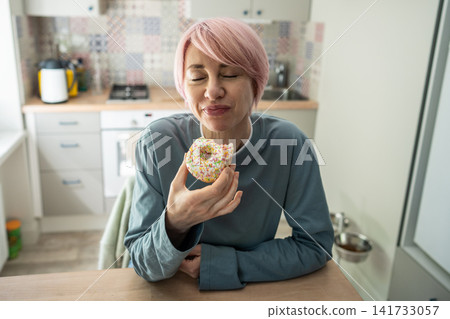 Pleased positive mature woman enjoying eating sweet glazed doughnut snack dessert in kitchen. Pleased positive mature woman enjoying eating sweet glazed doughnut snack dessert in kitchen. 141733057