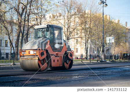 Road roller on street compacts freshly laid asphalt on traffic road, construction works in city. Road roller on street compacts freshly laid asphalt on traffic road, construction works in city. 141733112