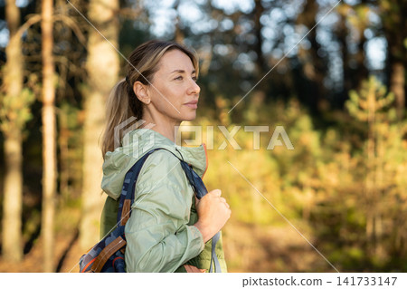 Confident positive woman hiking in forest with backpack, standing looking at sun enjoying quiet rest 141733147