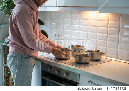 Man at home boiling cold water in pots on stove, temporary way to wash by heating water in stockpots Man at home boiling cold water in pots on stove, temporary way to wash by heating water in stockpots 141733186