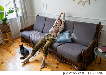 Sleepy bored woman stretching body on couch and yawning while spending lazy day at home with cat. 141733202