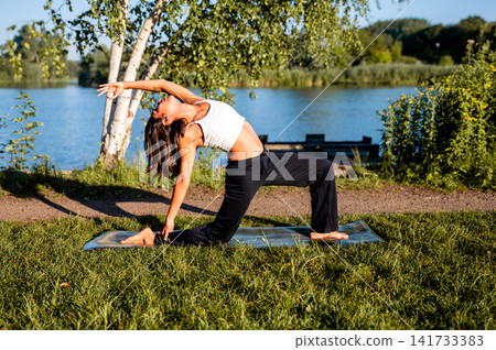 A young Hispanic woman practises a kneeling backbend surrounded by greenery in a park at golden hour, a mindful summer fitness and wellbeing practice outdoors. A young Hispanic woman practises a kneeling backbend surrounded by greenery in a park at golden hour, a mindful summer fitness and wellbeing practice outdoors. 141733383
