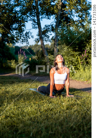 A young Hispanic woman practises the upward facing dog pose in a peaceful park by the lakeside in summer, connecting body and mind in nature in the tranquil evening. A young Hispanic woman practises the upward facing dog pose in a peaceful park by the lakeside in summer, connecting body and mind in nature in the tranquil evening. 141733386