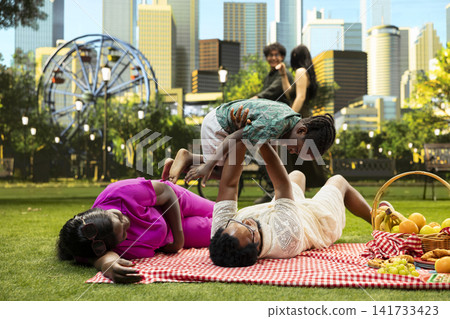 Father lifting child in airplane play during family picnic in park, parents laying on picnic blanket and playing with young boy beside basket of fruits and pastries. Father lifting child in airplane play during family picnic in park, parents laying on picnic blanket and playing with young boy beside basket of fruits and pastries. 141733423