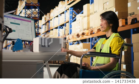 Asian man in wheelchair processing orders and preparing the boxes for distribution in fulfillment center, wears hi vis vest in e commerce logistics facility. Retail fast delivery. Camera A. Asian man in wheelchair processing orders and preparing the boxes for distribution in fulfillment center, wears hi vis vest in e commerce logistics facility. Retail fast delivery. Camera A. 141733436