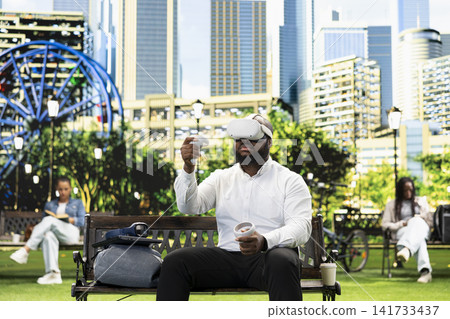 Businessman with virtual reality headset and motion controllers on park bench, with pedestrians in green background. VR glasses immersive simulation with tech. 141733437