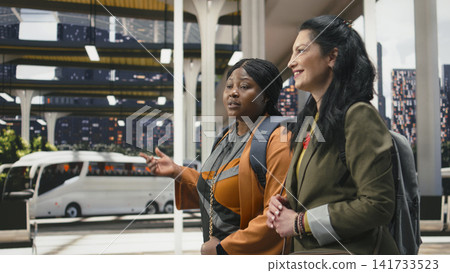 Smiling women travelers enjoying fun discussion as they move across coach station with backpacks on their shoulders. Joyous travel companions chatting and walking in bus terminal 141733523