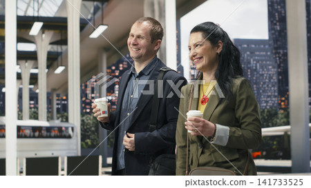 Smiling business travelers walk together through bus terminal with coffee cups in hand. Joyous businesspeople enjoying takeaway drinks in shuttle station, discussing travel itinerary Smiling business travelers walk together through bus terminal with coffee cups in hand. Joyous businesspeople enjoying takeaway drinks in shuttle station, discussing travel itinerary 141733525