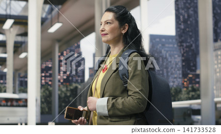 Smiling passenger holding mobile device while continuing through bus terminal. Jolly solo traveler checks digital route information on smartphone while walking in shuttle station Smiling passenger holding mobile device while continuing through bus terminal. Jolly solo traveler checks digital route information on smartphone while walking in shuttle station 141733528