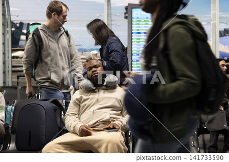Sleeping, neck pillow and tired man in airport waiting in lounge during delayed flights. Napping, headphones and exhausted african american person on chair in airport terminal during holiday trip Sleeping, neck pillow and tired man in airport waiting in lounge during delayed flights. Napping, headphones and exhausted african american person on chair in airport terminal during holiday trip 141733590
