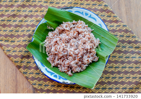 Close up of brown rice texture served on banana leaf. Healthy whole grain Asian food ingredient with natural daylight. Close up of brown rice texture served on banana leaf. Healthy whole grain Asian food ingredient with natural daylight. 141733920