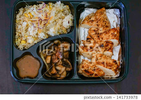 Top view of a grilled chicken breast meal served with rice, mushrooms and sauce in a compartmented takeaway box. Healthy, balanced protein food concept. Top view of a grilled chicken breast meal served with rice, mushrooms and sauce in a compartmented takeaway box. Healthy, balanced protein food concept. 141733978