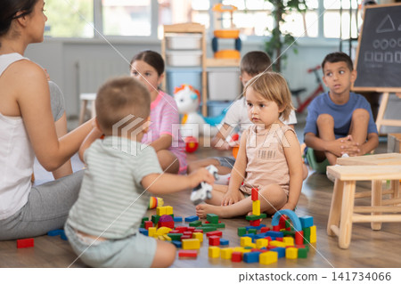 Caring young woman teacher playing educational games with group of children sitting on floor at daycare Caring young woman teacher playing educational games with group of children sitting on floor at daycare 141734066