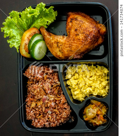 Top view of a roasted chicken leg bento meal served with rice, eggs and vegetables in a compartmented takeaway box. Balanced protein food concept. Top view of a roasted chicken leg bento meal served with rice, eggs and vegetables in a compartmented takeaway box. Balanced protein food concept. 141734874