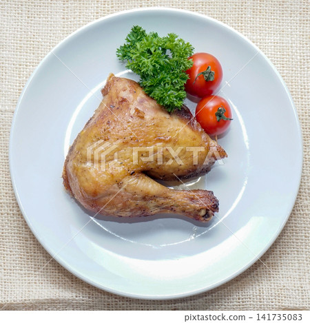 Top view of roasted chicken leg quarter served on a white plate with cherry tomatoes and parsley. Suitable for restaurant menus, food blogs, and editorial food content. 141735083