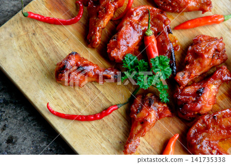 Top view of spicy chicken wings with red chili peppers on wooden board. Suitable for restaurant menus, BBQ promotions, food blogs, advertising and spicy cuisine concepts. Top view of spicy chicken wings with red chili peppers on wooden board. Suitable for restaurant menus, BBQ promotions, food blogs, advertising and spicy cuisine concepts. 141735338