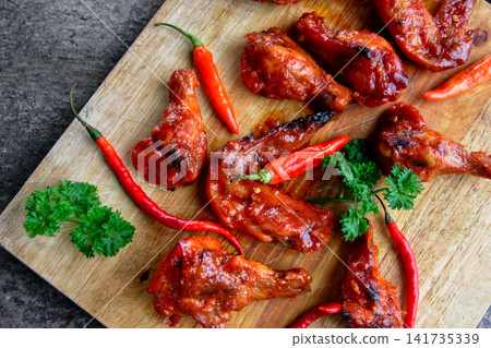 Top view of spicy chicken wings with red chili peppers on wooden board. Suitable for restaurant menus, BBQ promotions, food blogs, advertising and spicy cuisine concepts. Top view of spicy chicken wings with red chili peppers on wooden board. Suitable for restaurant menus, BBQ promotions, food blogs, advertising and spicy cuisine concepts. 141735339