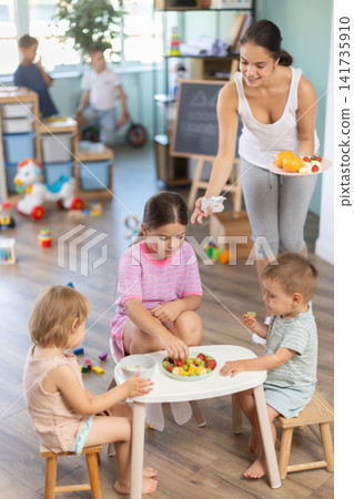 Children eat fruit at kindergarten. Light snack between games. Teacher serves fruit on tray Children eat fruit at kindergarten. Light snack between games. Teacher serves fruit on tray 141735910