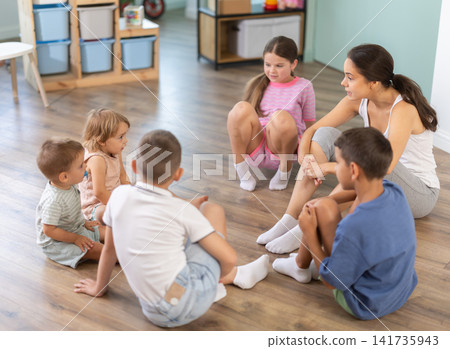 Children sitting on floor while caring female teacher explains lesson in kindergarten Children sitting on floor while caring female teacher explains lesson in kindergarten 141735943
