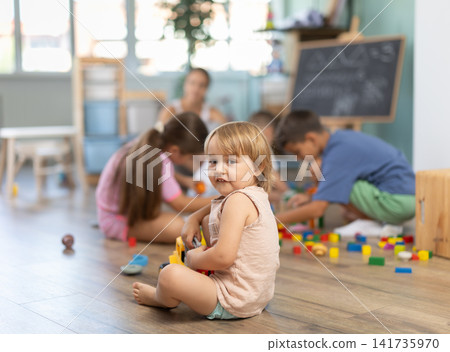 Baby boy sitting on floor and playing with cubes and cars in childcare center, other children in background Baby boy sitting on floor and playing with cubes and cars in childcare center, other children in background 141735970