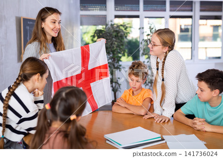 Schoolchildren listen carefully to story about the country England. Teacher holds flag of England in her hands Schoolchildren listen carefully to story about the country England. Teacher holds flag of England in her hands 141736024