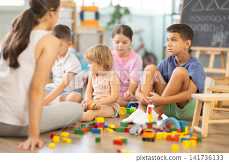 Attentive woman teacher sitting on floor together with small children playing educational games at classes for kids Attentive woman teacher sitting on floor together with small children playing educational games at classes for kids 141736133