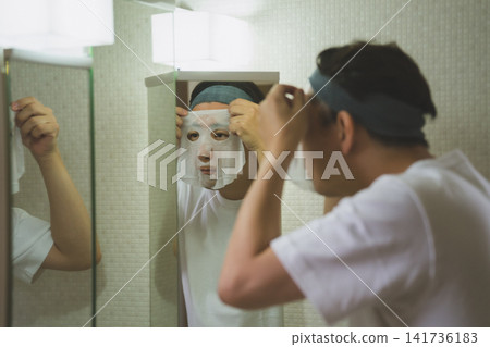 A 40-year-old man applying a sheet mask to his face in front of the bathroom mirror as part of his facial care routine. A 40-year-old man applying a sheet mask to his face in front of the bathroom mirror as part of his facial care routine. 141736183