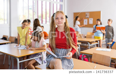 Portrait of schoolgirl with notebook in her hands who sits on desk in school class Portrait of schoolgirl with notebook in her hands who sits on desk in school class 141736337